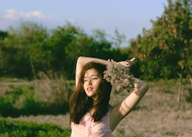 Asian woman in a pink dress posing joyfully with a bouquet in a sunny outdoor field.