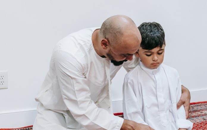 A father kneels with his son in prayer, both wearing white robes, against a backdrop of string lights.